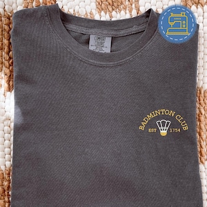 Może przedstawiać: Ciemnoszara koszulka z napisem "Badminton Club" i żółtą grafiką lotki. Widnieje również napis "EST 1754". Koszulka wykonana jest z miękkiego materiału.