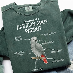 May include: A dark green sweatshirt featuring a detailed illustration of an African Grey Parrot. The graphic includes labeled anatomical features like "Wisdom Processor" and "Mimic Speaker." The text "Anatomy of a African Grey Parrot" is printed above the bird.