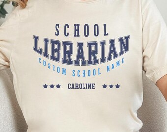 Puede incluir: Camiseta color crema con el texto "SCHOOL LIBRARIAN" en azul marino en forma de arco. Debajo, dice "CUSTOM SCHOOL NAME" y "CAROLINE" con tres estrellas a cada lado. La camiseta es de color crema suave.