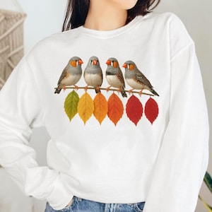 Puede incluir: Sudadera blanca con un gráfico de cuatro pájaros pequeños posados en una rama con hojas de otoño coloridas. Los pájaros tienen plumas grises y marrones con picos naranjas. Las hojas son verdes, amarillas, naranjas y rojas.