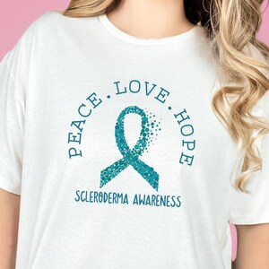 Könnte beinhalten: Weißes T-Shirt mit einem Design aus türkisfarbenem Band. Das Band besteht aus kleinen Kreisen und trägt die Worte "Peace. Love. Hope" darum herum. Der Text "Scleroderma Awareness" befindet sich unterhalb des Bandes.