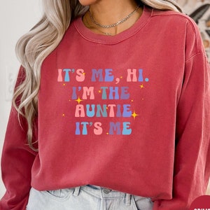 Könnte beinhalten: Ein roter Pullover mit dem Text "It's me, hi. I'm the auntie. It's me" in einer bunten Retro-Schrift mit Sternen.