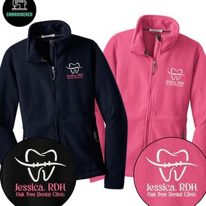 May include: Navy blue and pink fleece zip-up jackets. Each jacket features an embroidered dental logo with the text "Jessica, RDH Oak Tree Dental Clinic". The jackets have a high collar and a zipper closure.