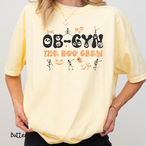 May include: Butter yellow t-shirt with the text "OB-GYN THE BOO CREW" in black and orange. The design includes Halloween-themed graphics such as skeletons, ghosts, bats, and jack-o'-lanterns. The shirt has short sleeves and a crew neck.