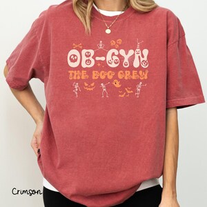 May include: Crimson-colored t-shirt with the text "OB-GYN THE BOO CREW" in a Halloween-themed design. The design includes skulls, skeletons, ghosts, and bats in orange and white. The shirt is a short-sleeved, crew neck style.