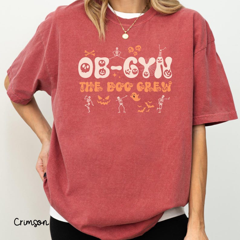 May include: Crimson-colored t-shirt with the text "OB-GYN THE BOO CREW" in a Halloween-themed design. The design includes skulls, skeletons, ghosts, and bats in orange and white. The shirt is a short-sleeved, crew neck style.