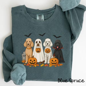Puede incluir: Sudadera azul abeto con un diseño de Halloween. El gráfico muestra cuatro caniches con disfraces de Halloween, incluyendo un fantasma, una momia y un vampiro, con calabazas y murciélagos. La sudadera tiene una etiqueta Comfort Colors.