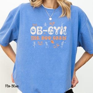 May include: Flo Blue sweatshirt with the text "OB-GYN THE BOO CREW" in orange. The design includes a skull, ghosts, skeletons, and bats. The sweatshirt is a solid light blue color.
