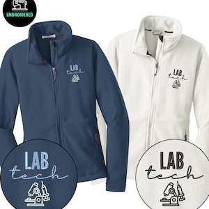 May include: Two fleece jackets, one navy blue and one white, with a full zipper and a high collar. Each jacket has the embroidered text "LAB TECH" with a microscope and syringe design. The jackets are designed for lab professionals.