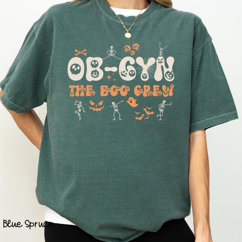 May include: A teal green t-shirt with the text "OB-GYN THE BOO CREW" in orange and white. The shirt features Halloween-themed graphics, including skulls, skeletons, ghosts, and bats. The shirt is a comfortable, casual style.