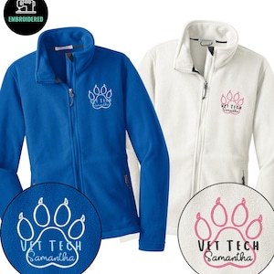 Könnte beinhalten: Zwei Fleecejacken, eine blaue und eine weiße, mit einem Pfotenabdruck-Design und dem Text "VET TECH Samantha" auf der Brust gestickt. Die Jacken haben einen durchgehenden Reißverschluss und einen hohen Kragen. Das Bild zeigt auch eine Nahaufnahme der Stickerei.
