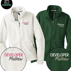 May include: Two fleece zip-up jackets, one white and one dark green. Each jacket features the embroidered text "DEVELOPER Mathew" on the chest. The jackets have a high collar and a full-length zip. The top left corner has a black circle with the word "EMBROIDERED".