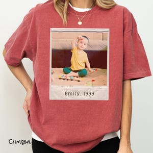 Peut inclure: T-shirt couleur cramoisi avec un col blanc. Le t-shirt pr&eacute;sente une photographie encadr&eacute;e d'un enfant en chemise jaune et pantalon turquoise. La photographie est l&eacute;gend&eacute;e "Emily, 1999."