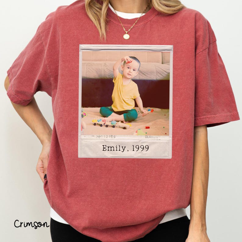 Peut inclure: T-shirt couleur cramoisi avec un col blanc. Le t-shirt pr&eacute;sente une photographie encadr&eacute;e d'un enfant en chemise jaune et pantalon turquoise. La photographie est l&eacute;gend&eacute;e "Emily, 1999."
