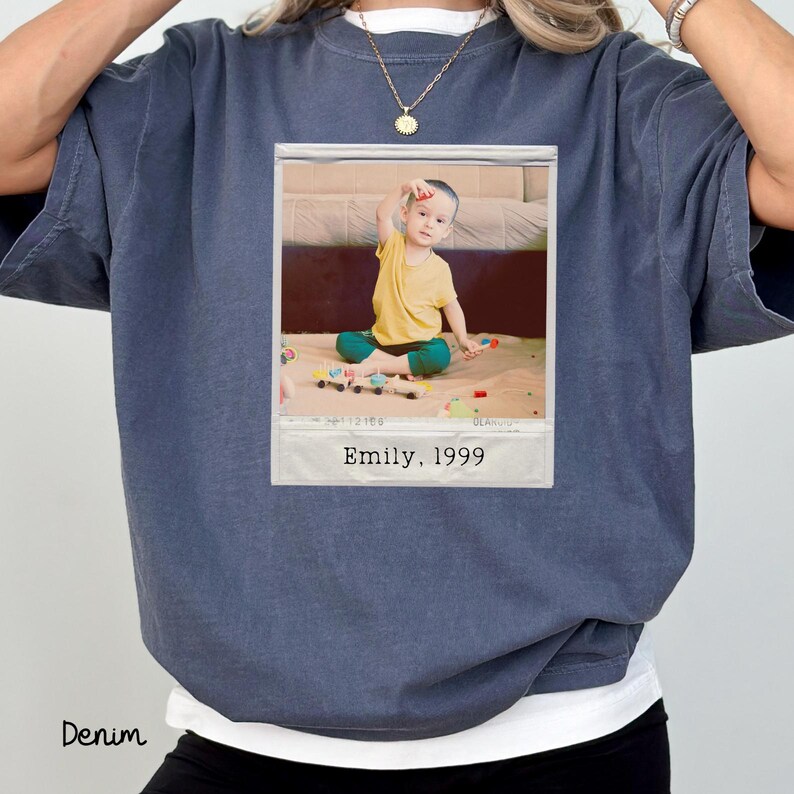 Peut inclure: T-shirt bleu denim avec une impression d'une photo polaro&iuml;d d'un enfant en chemise jaune et pantalon vert. La photo porte l'inscription "Emily, 1999". Le mot "Denim" est imprim&eacute; en bas &agrave; gauche.