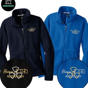 May include: Two fleece zip-up jackets, one navy blue and one bright blue. Each jacket has the embroidered text "Alexa B(R) Radiology" in gold thread on the chest. The jackets have a high collar and a full-length zipper.