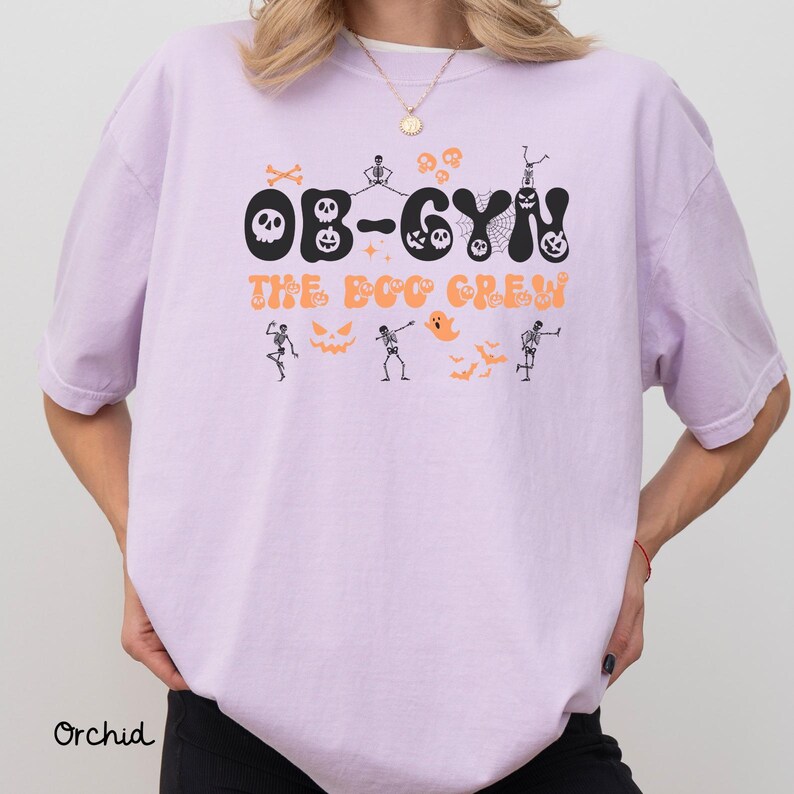 May include: Orchid-colored t-shirt with the text "OB-GYN The Boo Crew" in black and orange. The design includes Halloween-themed graphics such as skeletons, skulls, and bats. The shirt is a comfortable, casual style.