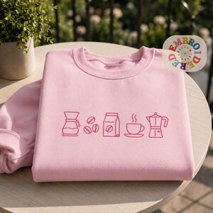 Puede incluir: Una sudadera de cuello redondo en color rosa claro con un diseño bordado de arte lineal en rosa oscuro en el pecho. El gráfico muestra una jarra de café, tres granos de café, una bolsa de café, una taza de café humeante y una cafetera moka.