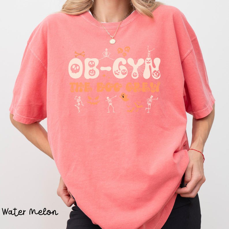 May include: A watermelon-colored t-shirt with the text "OB-GYN The Boo Crew" in a Halloween-themed design. The design includes skeleton figures, pumpkins, and ghosts. The shirt is a comfortable, casual style.