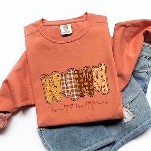 Puede incluir: Una sudadera de color coral con la palabra "NANA" en un diseño con temática otoñal. Las letras están rellenas con patrones de hojas y cuadros. Debajo de la palabra están los nombres "Sophia", "Ryan" y "Emily".