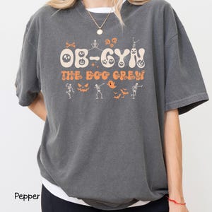 May include: A dark gray t-shirt with the text "OB-GYN THE BOO CREW" in orange and white. The design includes Halloween-themed graphics such as skeletons, bats, and jack-o'-lanterns. The shirt is a comfortable, oversized fit.