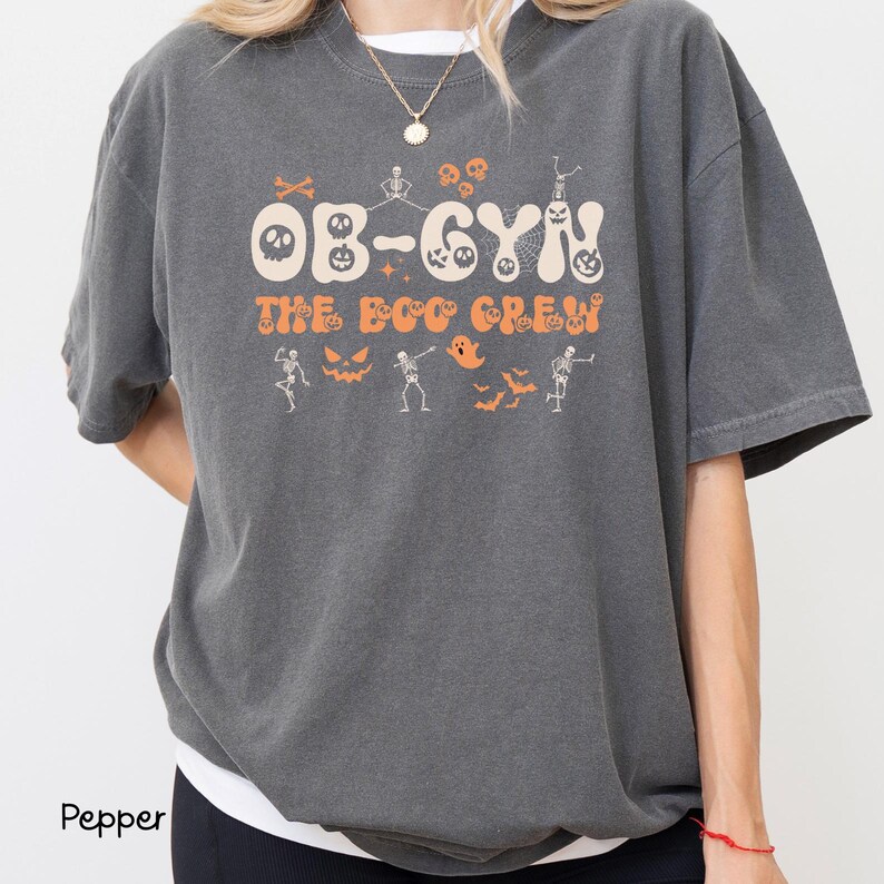 May include: A dark gray t-shirt with the text "OB-GYN THE BOO CREW" in orange and white. The design includes Halloween-themed graphics such as skeletons, bats, and jack-o'-lanterns. The shirt is a comfortable, oversized fit.