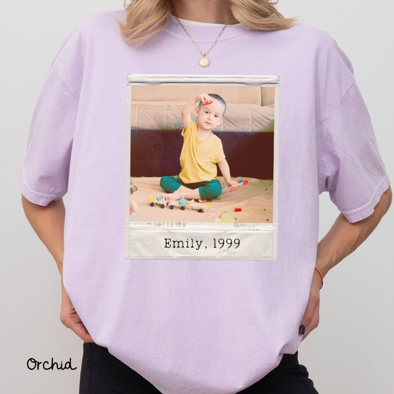 Peut inclure: T-shirt de couleur orchid&eacute;e avec une photographie encadr&eacute;e d'un enfant. La photo montre un enfant en chemise jaune et pantalon vert jouant avec des jouets. Le texte "Emily, 1999" est imprim&eacute; sur le cadre.