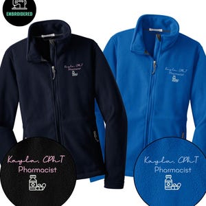 Puede incluir: Dos chaquetas de forro polar con cremallera, una azul marino y otra azul real. Cada chaqueta tiene el nombre "Kayla, CPhT, Pharmacist" bordado en rosa. Las chaquetas tienen cuello alto y cierre de cremallera.