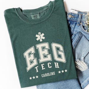 Può includere: Una maglietta verde con una grafica bianca che dice "EEG Tech" con un simbolo di stella della vita sopra il testo e tre stelle sotto il testo. Il testo "CAROLINE" è sotto le stelle.