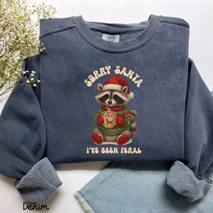 Könnte beinhalten: Ein denimblaues Sweatshirt mit dem Text "Sorry Santa, I've Been Feral". Die Grafik zeigt einen Waschbären mit Weihnachtsmütze und Weihnachtspullover, der eine Tasse hält. Das Sweatshirt hat einen Rundhalsausschnitt und lange Ärmel.