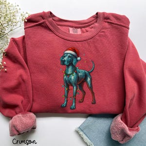 May include: Crimson-colored sweatshirt featuring a blue dog illustration wearing a Santa hat and Christmas lights. The dog has a mosaic-like design. The text "Crimson" is at the bottom of the sweatshirt.