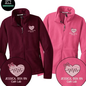 May include: Two fleece zip-up jackets, one burgundy and one pink. Each jacket features an embroidered heart design with a bow and a heartbeat line, along with the text "JESSICA, BSN RN Cath Lab". The jackets have a high collar and a zipper closure.
