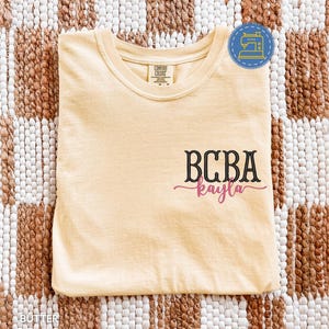 Puede incluir: Camiseta de color mantequilla con el texto "BCBA kayla" en escritura negra y rosa. La camiseta tiene cuello redondo y mangas cortas. La etiqueta de la marca "Comfort Colors" es visible.