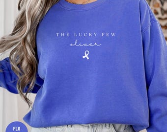 May include: A royal blue sweatshirt featuring the white text "THE LUCKY FEW oliver" and a white ribbon graphic. The sweatshirt has a crew neck and long sleeves. A small circle in the bottom left corner reads "FLO BLUE".
