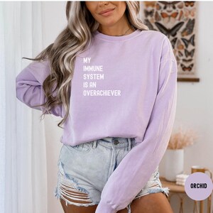 Peut inclure: Un sweat-shirt lilas avec le texte blanc "MY IMMUNE SYSTEM IS AN OVERACHIEVER". Le haut à manches longues est de couleur unie. Le modèle porte un short en jean.