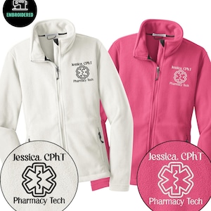 May include: Two fleece jackets, one white and one pink, with a full zipper and a stand-up collar. Each jacket has the embroidered text "Jessica, CPhT Pharmacy Tech" and a medical symbol. The jackets are designed for pharmacy technicians.