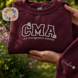 May include: A maroon crewneck sweatshirt featuring white embroidered text that reads CMA and case management assistant, accented with small white daisy flowers. The sleeve includes the embroidered name Kayla with a small daisy flower.