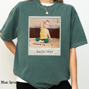 Peut inclure: Un t-shirt vert d'eau avec une photographie encadr&eacute;e d'un enfant en chemise jaune et pantalon vert. La photographie est l&eacute;gend&eacute;e "Emily, 1999". Le t-shirt a une coupe ample et des manches courtes. Les mots "Blue Spruce" sont imprim&eacute;s en bas &agrave; gauche.