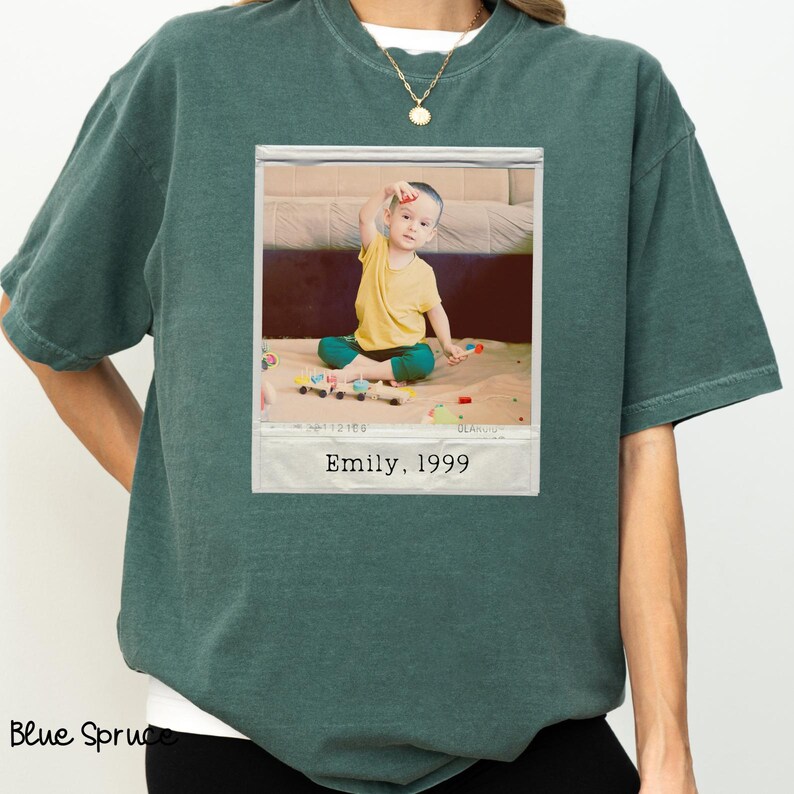 Peut inclure: Un t-shirt vert d'eau avec une photographie encadr&eacute;e d'un enfant en chemise jaune et pantalon vert. La photographie est l&eacute;gend&eacute;e "Emily, 1999". Le t-shirt a une coupe ample et des manches courtes. Les mots "Blue Spruce" sont imprim&eacute;s en bas &agrave; gauche.