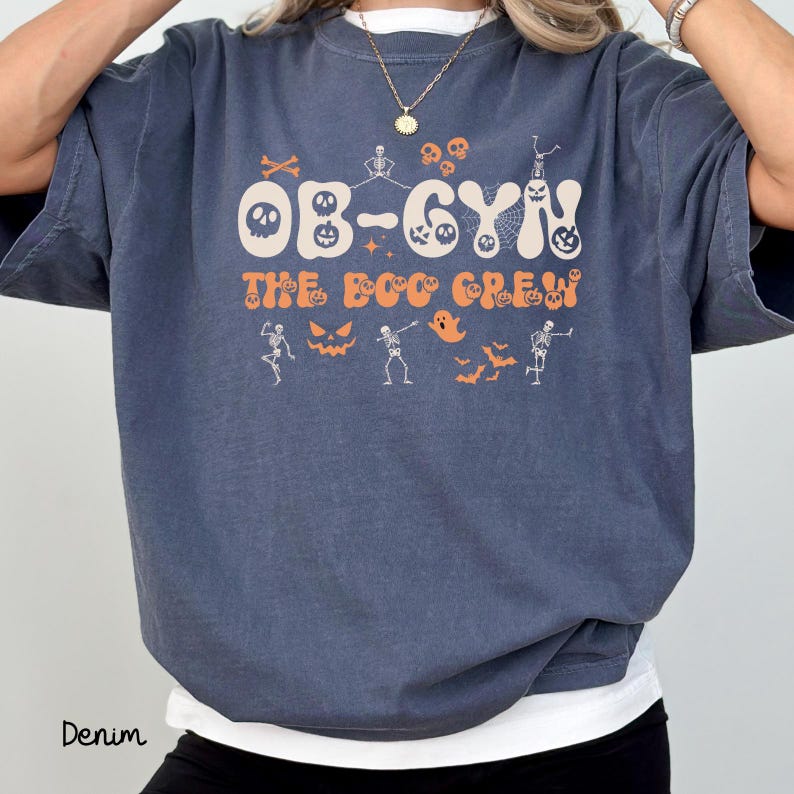 May include: Denim blue t-shirt with the text "OB-GYN The Boo Crew" in orange. The shirt features Halloween-themed graphics, including skeletons, bats, and jack-o'-lanterns. The shirt is a relaxed fit.