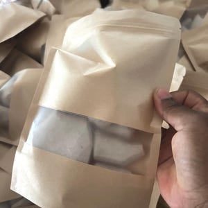 May include: A light brown paper stand-up pouch with a clear rectangular plastic window on the front. The pouch features a resealable zipper top and contains several irregular, opaque white solid pieces visible through the transparent section.