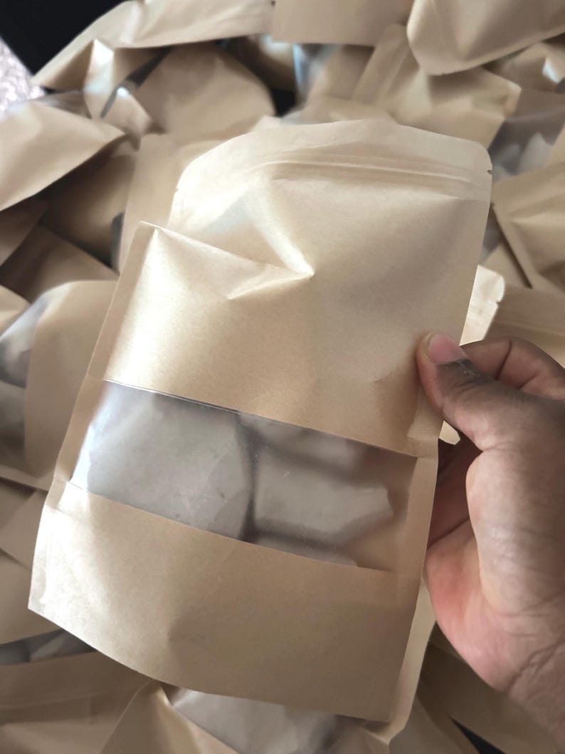 May include: A light brown paper stand-up pouch with a clear rectangular plastic window on the front. The pouch features a resealable zipper top and contains several irregular, opaque white solid pieces visible through the transparent section.