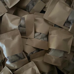 May include: A collection of multiple light brown kraft paper pouches arranged in a pile. Each pouch features a rectangular transparent plastic window on the front, allowing the contents inside to be partially visible through the clear material.