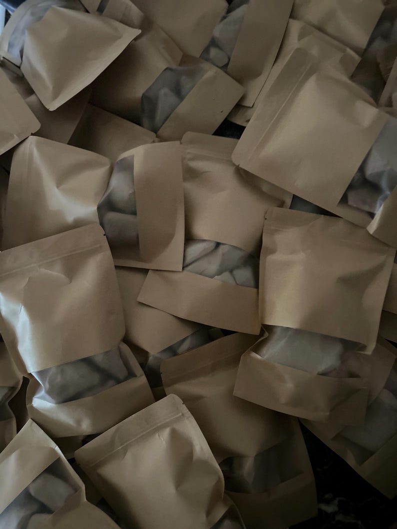 May include: A collection of multiple light brown kraft paper pouches arranged in a pile. Each pouch features a rectangular transparent plastic window on the front, allowing the contents inside to be partially visible through the clear material.