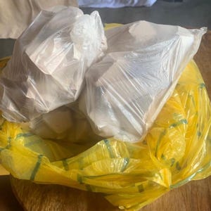 May include: Two clear, thin plastic bags filled with white, irregularly shaped solid pieces are nested inside a larger, translucent yellow plastic bag. The bags are gathered at the top, showing a crinkled texture and semi-transparent appearance.
