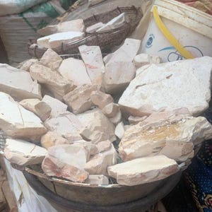 May include: A large collection of irregular, off-white and beige mineral chunks piled inside a circular metal wire basket. The stones have a matte, chalky texture with visible jagged edges and varying sizes, displaying natural earthy tones throughout.