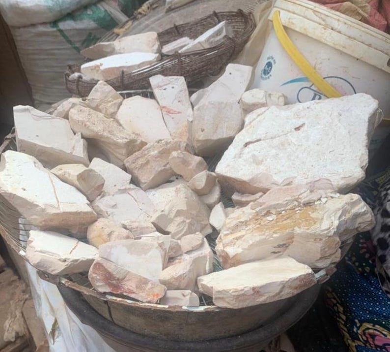 May include: A large collection of irregular, off-white and beige mineral chunks piled inside a circular metal wire basket. The stones have a matte, chalky texture with visible jagged edges and varying sizes, displaying natural earthy tones throughout.