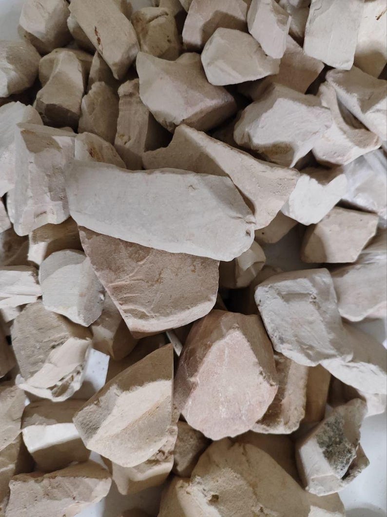 May include: A collection of irregular, raw clay chunks in shades of off-white, cream, and pale beige. These solid, earthy fragments feature matte, chalky textures with sharp, jagged edges and uneven surfaces, piled together in a dense, compact cluster.