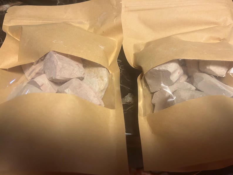 May include: Two light brown paper pouches with clear plastic windows, each containing several irregular, off-white, chalky rock fragments. The pouches are sealed at the top and have a matte texture, displaying the solid mineral pieces inside.