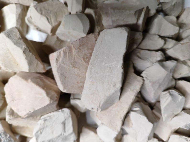 May include: Close-up view of a pile of off-white and light beige clay pieces. The irregular shapes and sizes of the clay chunks are clearly visible, showcasing their rough, naturally broken textures.  The color palette is consistent throughout, with subtle variations in shade. The image highlights the raw, unrefined nature of the clay material, suitable for pottery, sculpting, or other crafting projects.